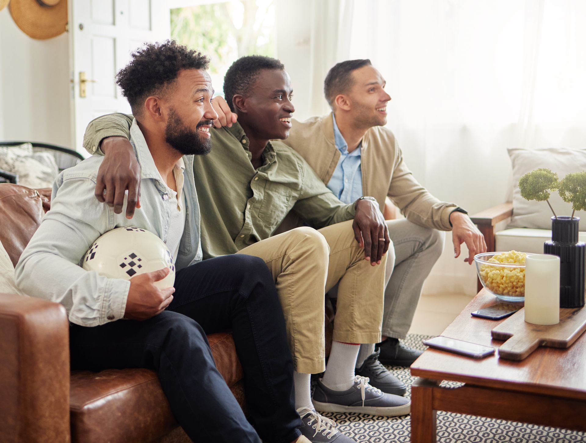Three men on a couch watching TV. One holds a soccer ball. They are smiling, indoors.