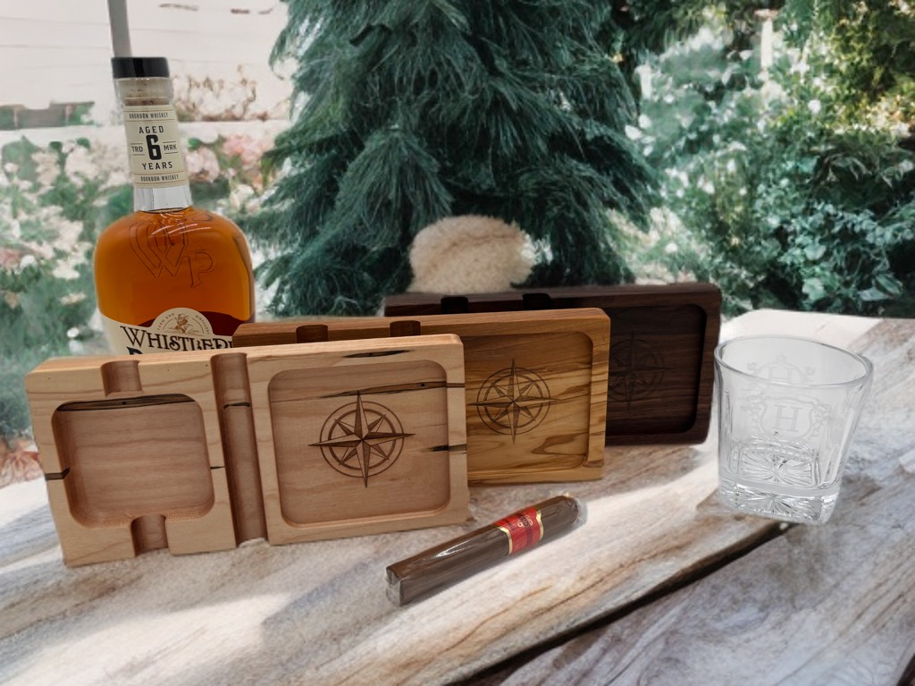Wooden boxes with compass designs, bourbon bottle, cigar, and a glass on a rustic surface.