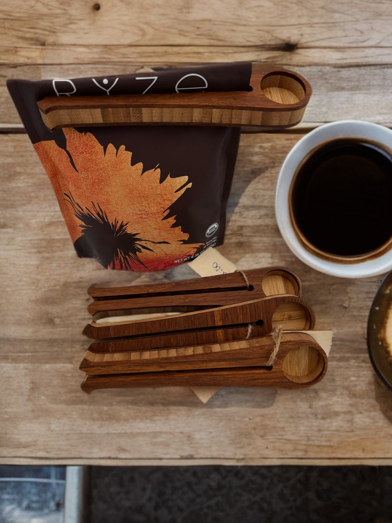 Wooden coffee scoopers with a coffee bag and a cup of coffee on a wooden table.