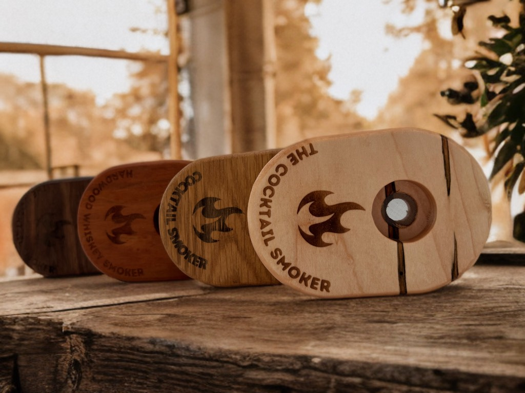 Wooden cocktail smokers in various wood tones, with logo and hole, lined up on a rustic surface outdoors.
