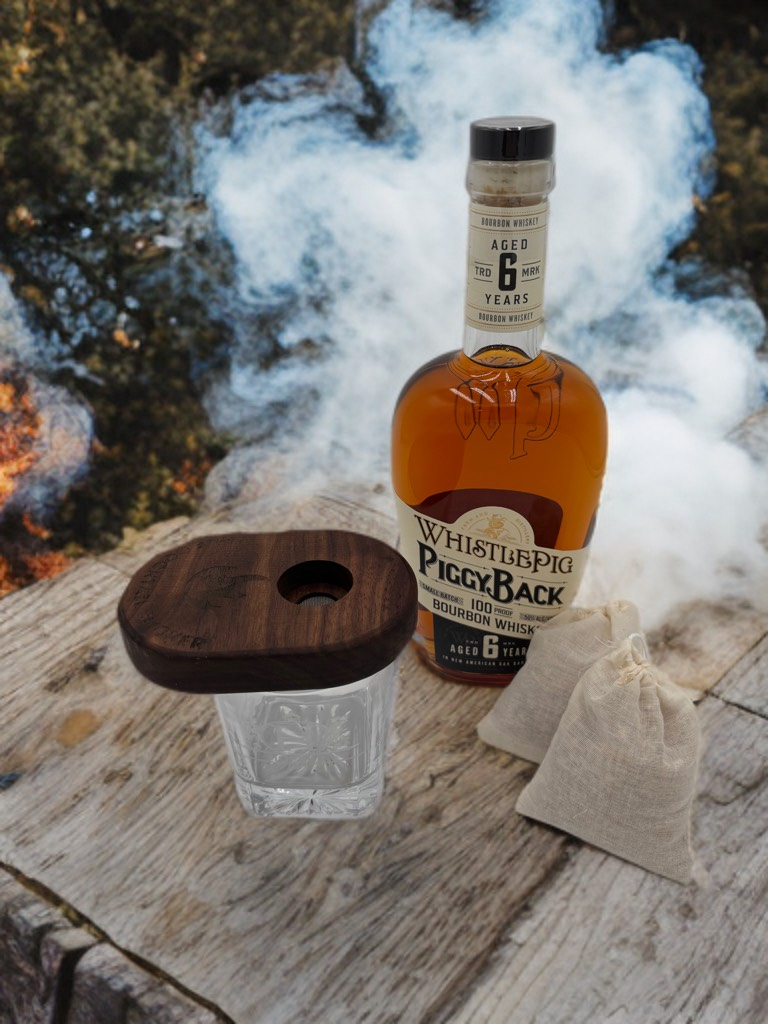 Bourbon bottle, glass, wooden lid, and two small bags on a wooden surface with smoke in background.