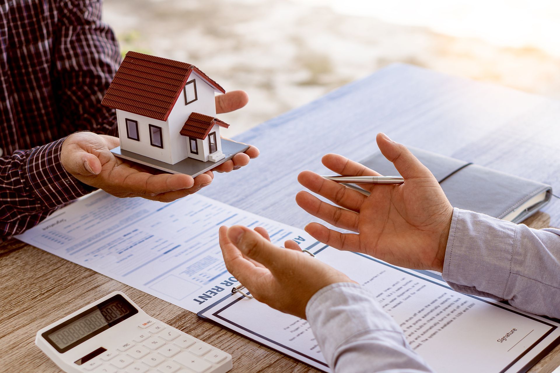 Hands exchanging house keys over real estate papers with a small model home and calculator on a table