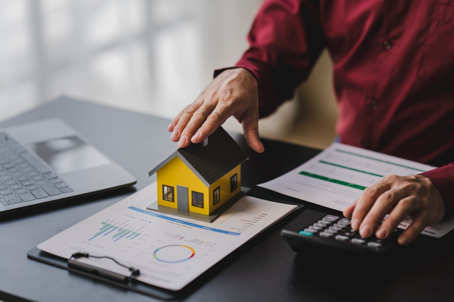 Hands on a house model beside charts, calculator, and laptop on a desk