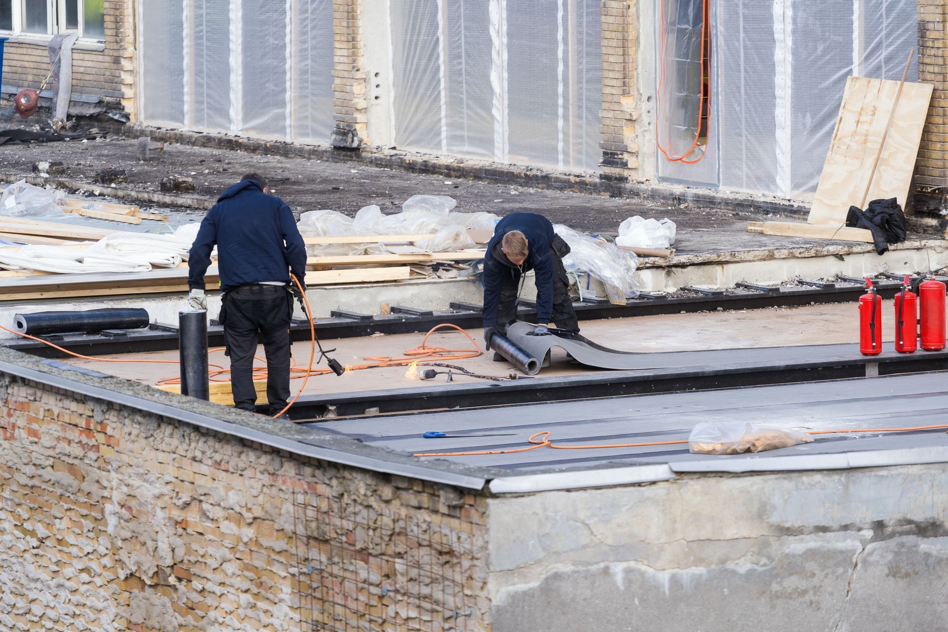 Two workers installing a flat roof, using a torch and rolls of roofing material on a building.
