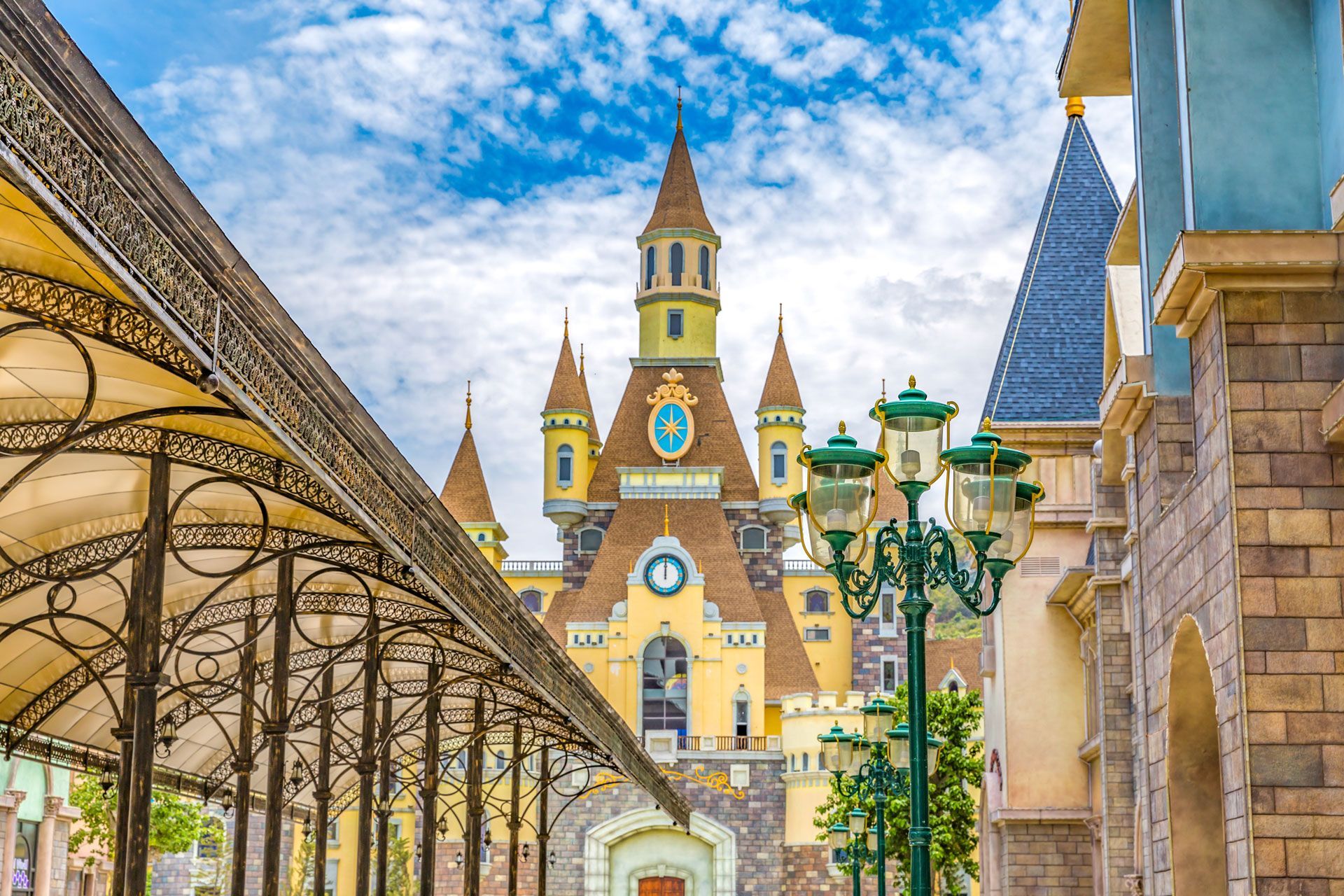 A pale yellow fairytale castle with blue-faced clocks and ornate turrets under a bright, cloudy blue sky.