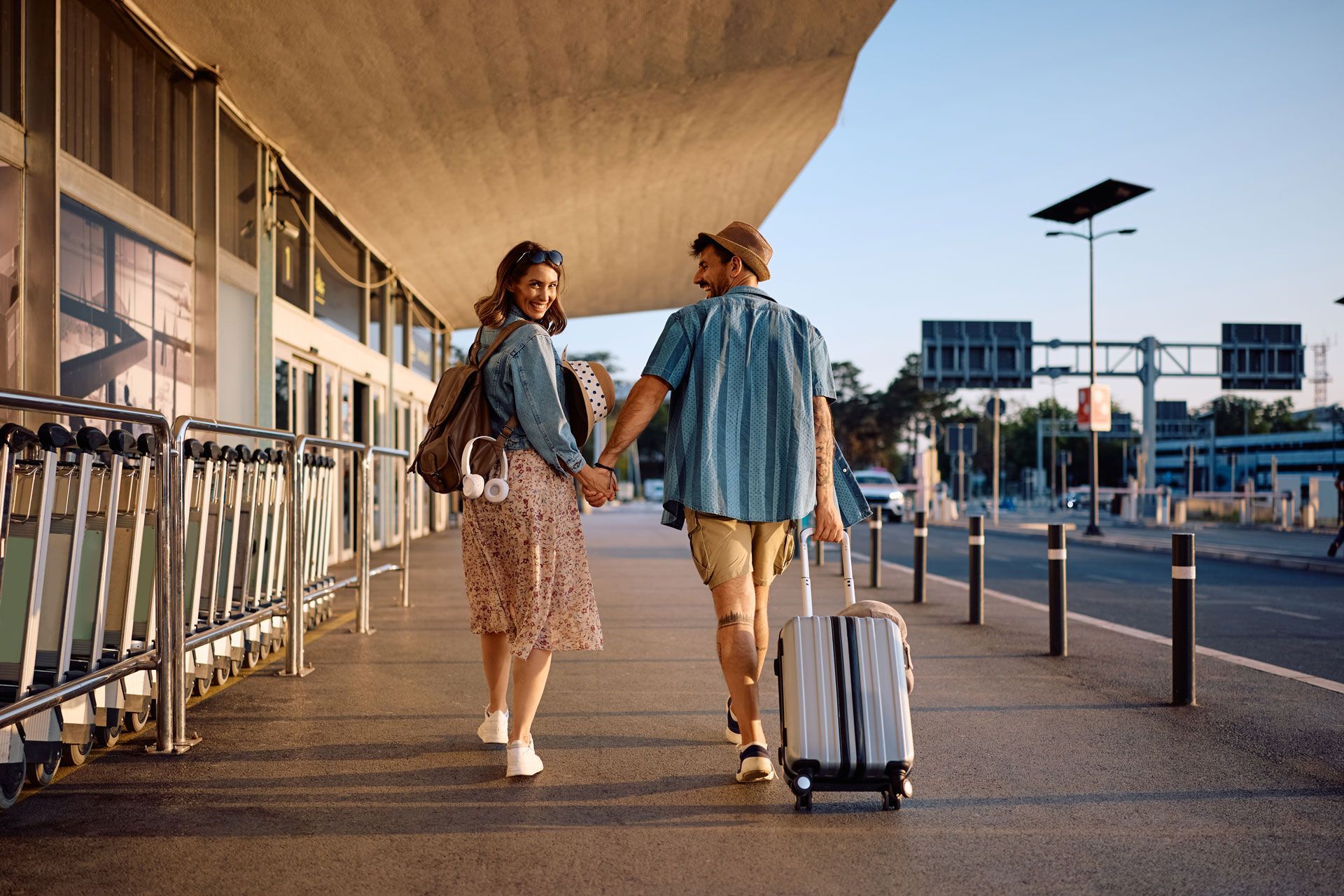 A couple walks hand-in-hand with luggage outside an airport terminal during a sunny day.