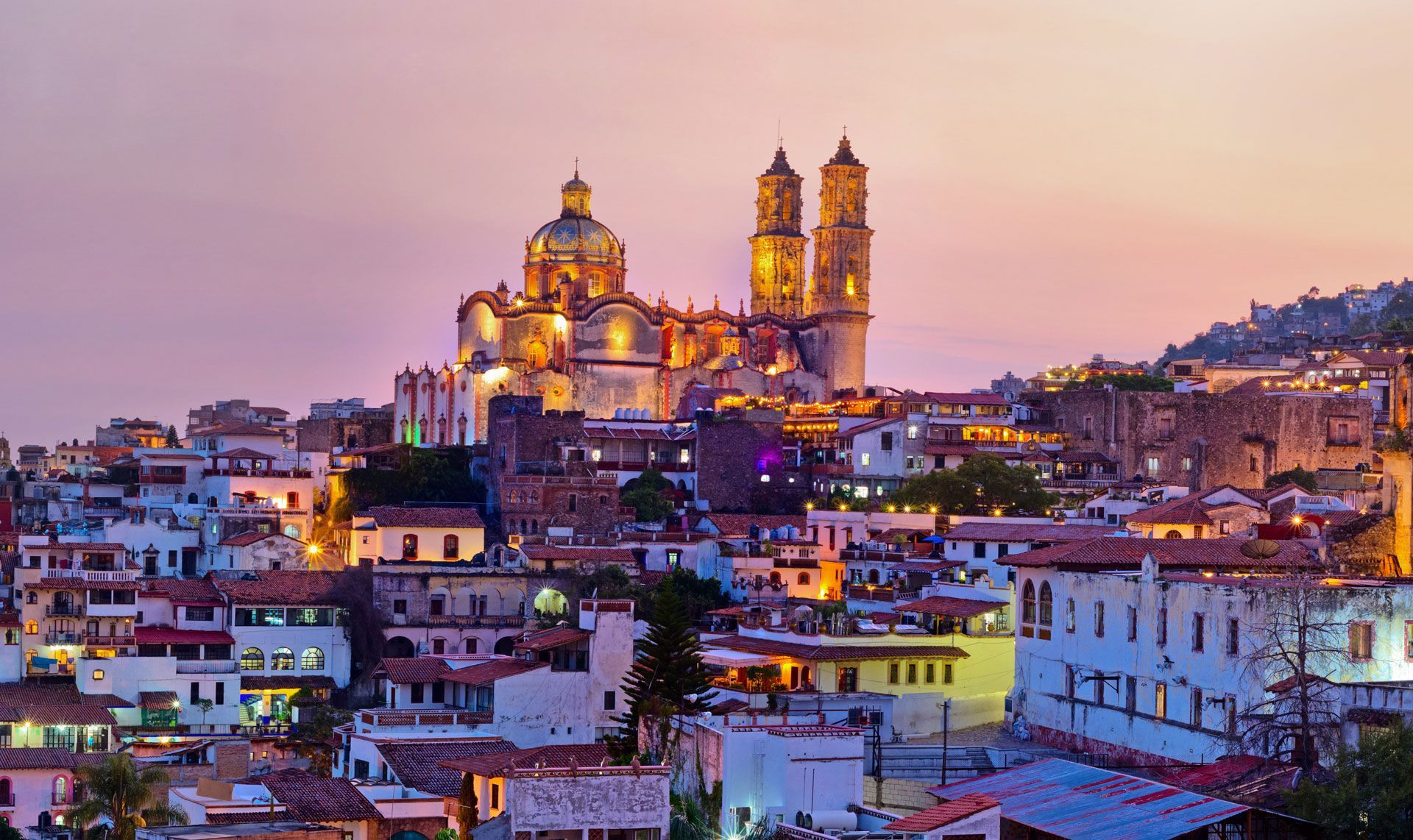 A warm, sunset-lit view of Taxco, Mexico, featuring the historic Santa Prisca Church rising above white-walled buildings.