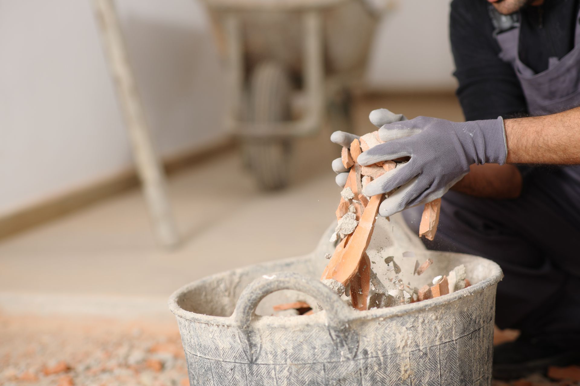 A worker wearing grey gloves places pieces of broken brick into a plastic bucket at a construction site.