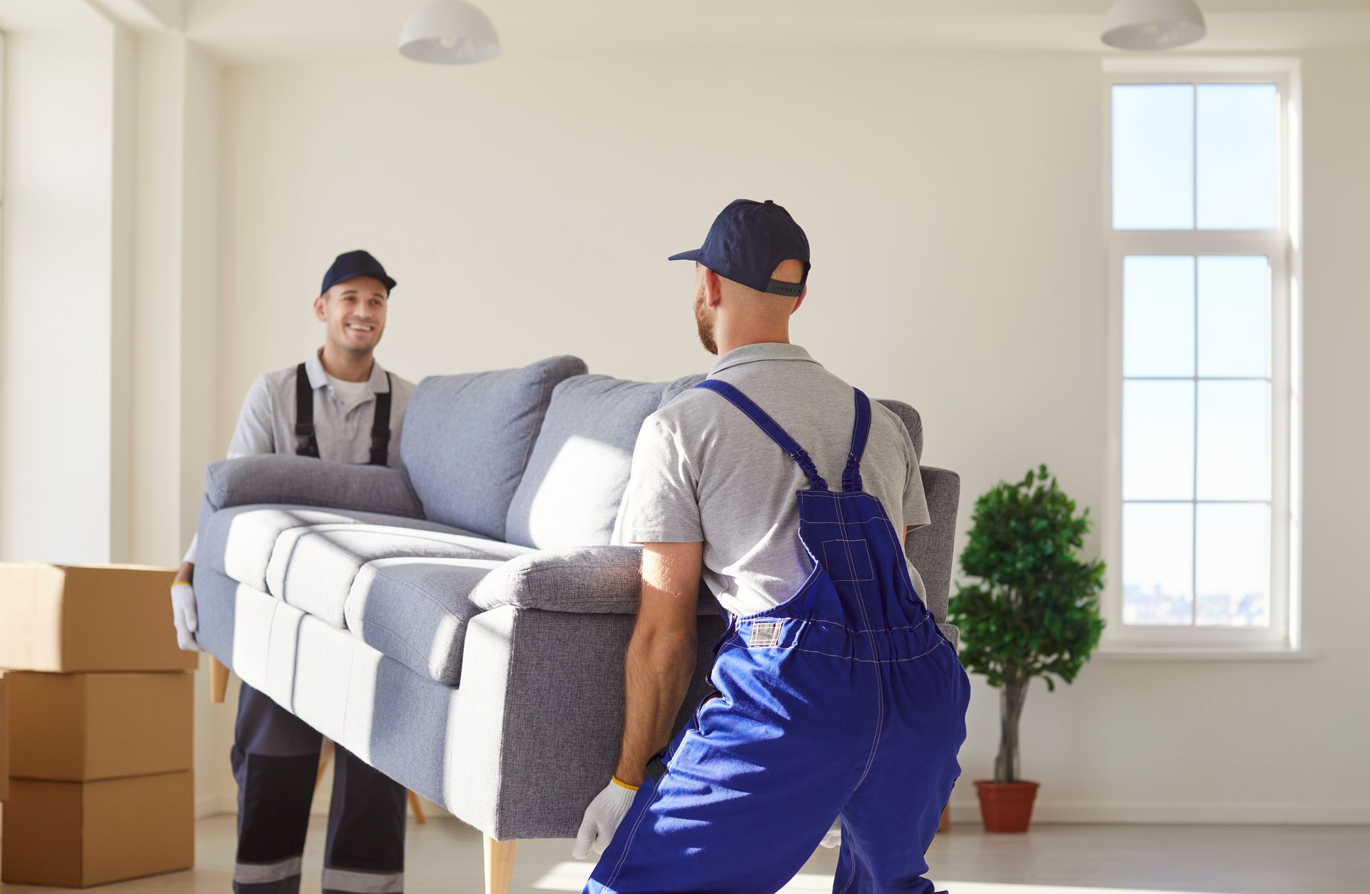 Two movers in blue work uniforms carrying a grey sofa across a bright, empty room with cardboard boxes and a small plant.
