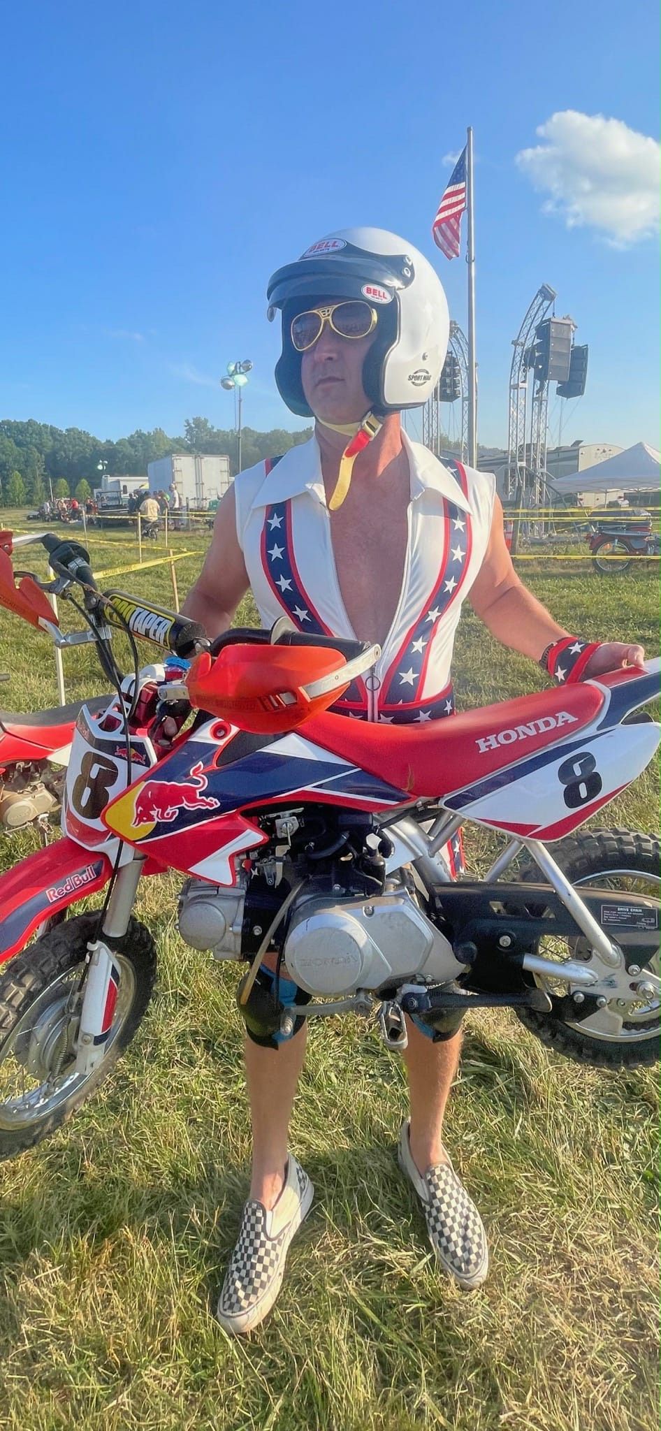Man in patriotic outfit and helmet poses with a red dirt bike outdoors, American flag in background.