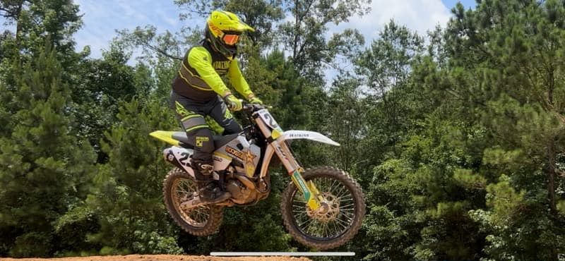 Motocross rider in yellow gear jumps a dirt track ramp with trees in the background.