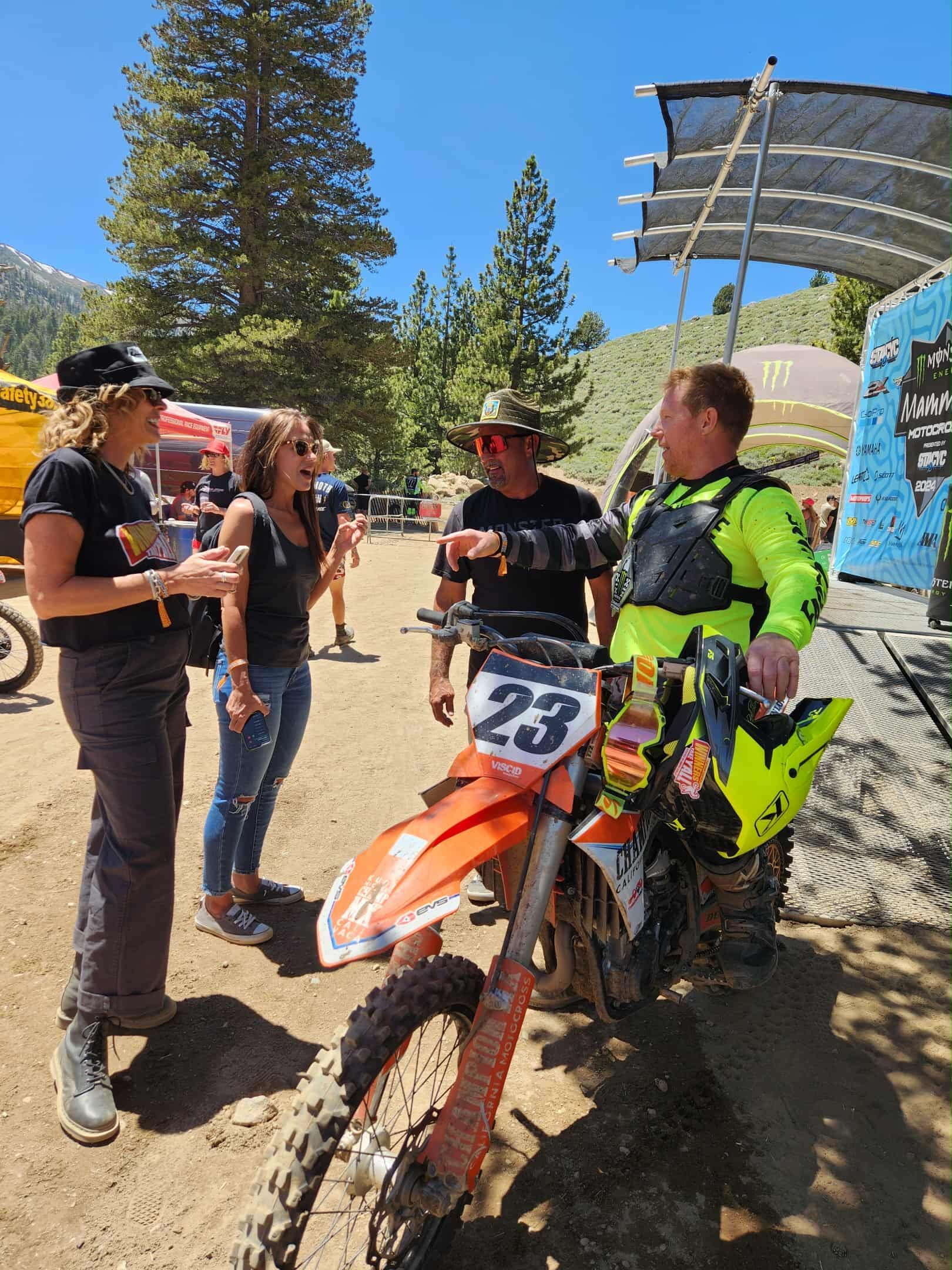 People converse near a dirt bike with the number 23. Sunny outdoor setting.