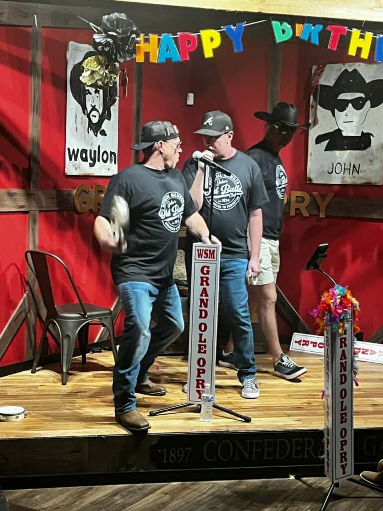 Three men on stage with microphones in front of a “Grand Ole Opry” backdrop, one holding a tambourine.