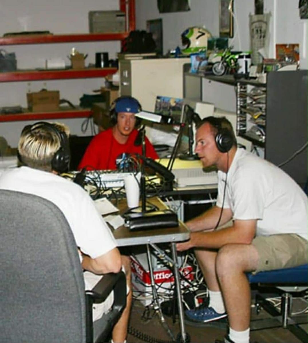Three people wearing headphones at a table, recording in a cluttered room with shelves and equipment.