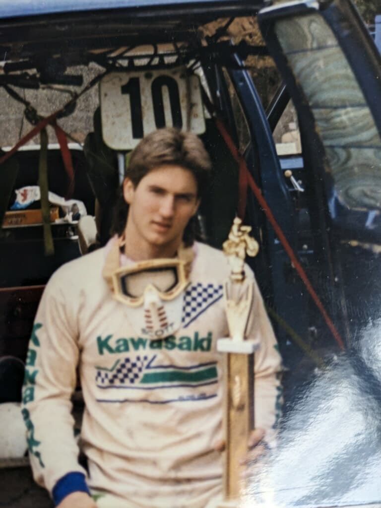 Man in Kawasaki racing gear, holding a trophy, standing in front of a race car.