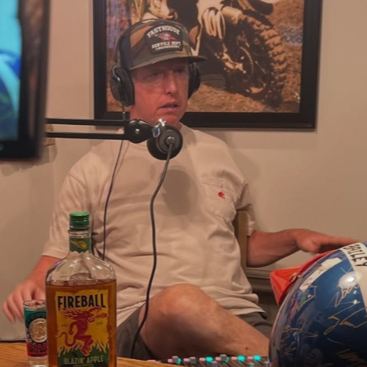 Man wearing headphones sits, speaking into a microphone. A bottle of Fireball whiskey sits on a table.