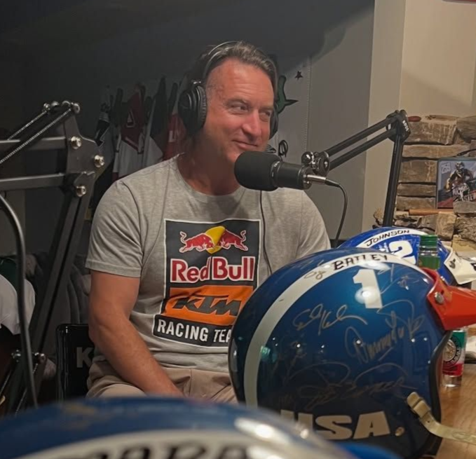 Man wearing headphones and Red Bull Racing tee, speaking into a microphone in a studio setting. Blue helmets are nearby.