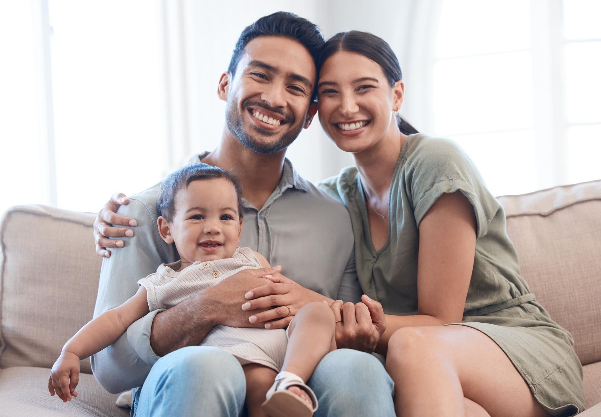 A smiling family of three sits on a couch indoors.