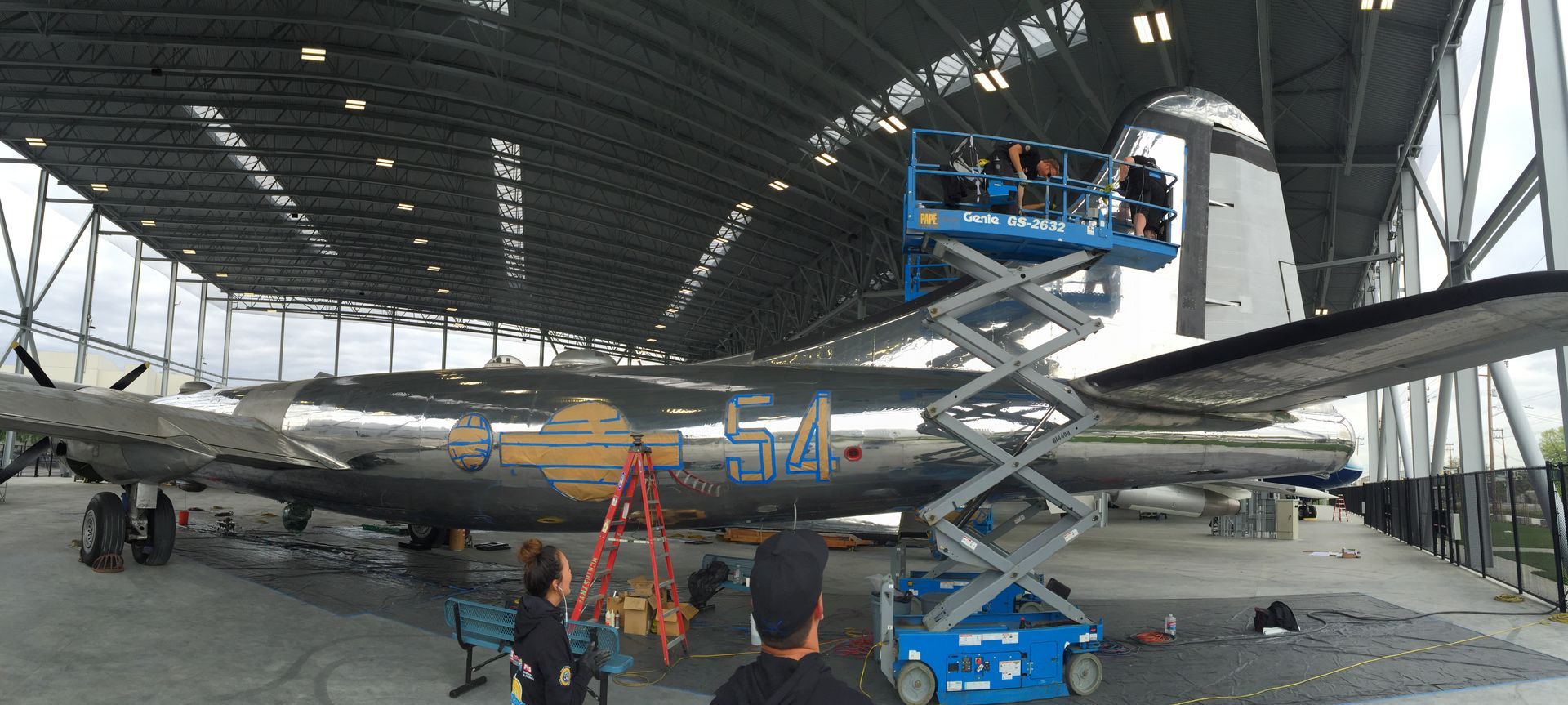 People use a scissor lift and ladder to restore a vintage, metallic airplane inside a large, open aircraft hangar.