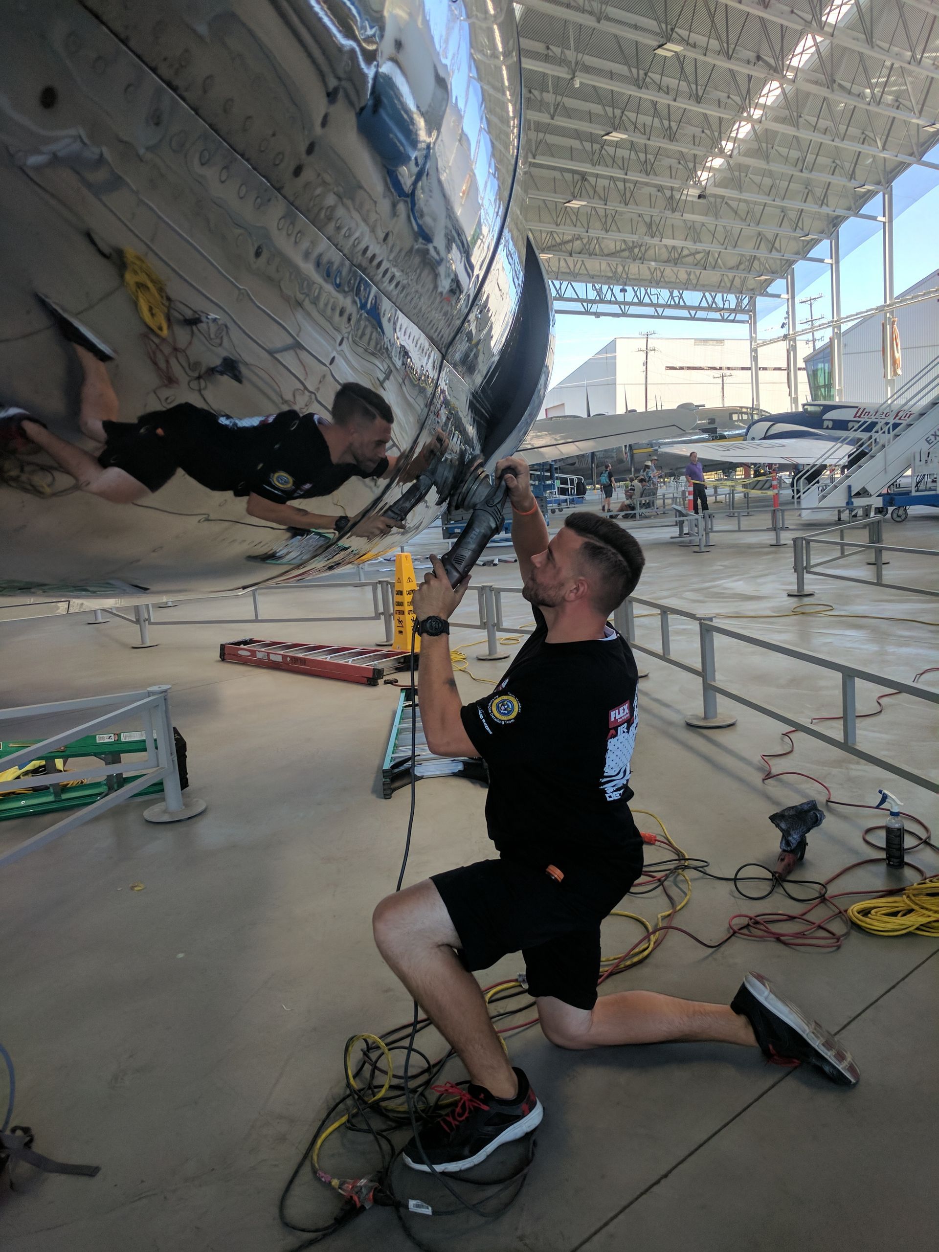 A person kneels while polishing the reflective surface of an aircraft in a large, open-air hangar.