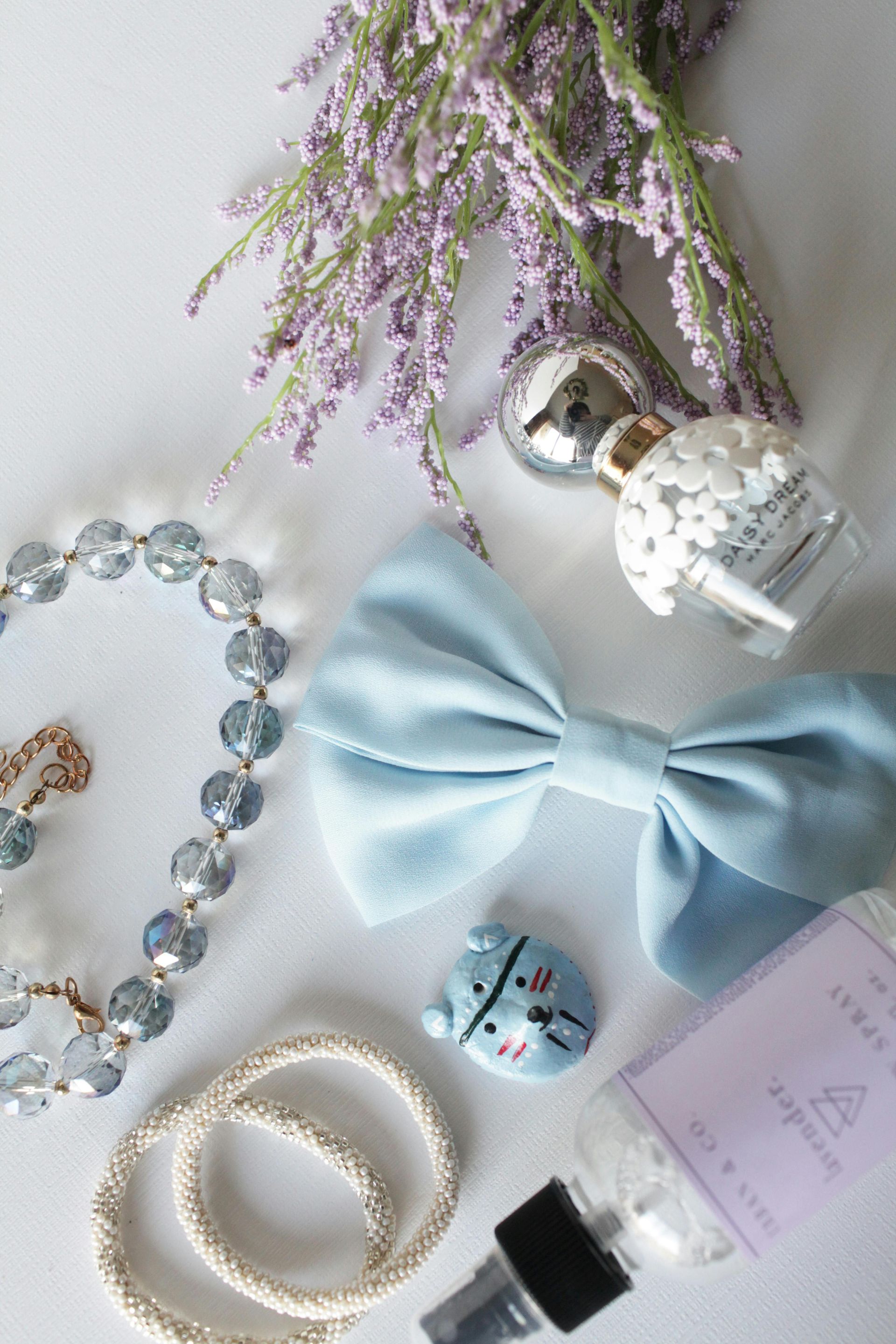 Still life: light blue bow, jewelry, lavender sprigs, perfume bottle, and small trinkets on white background.