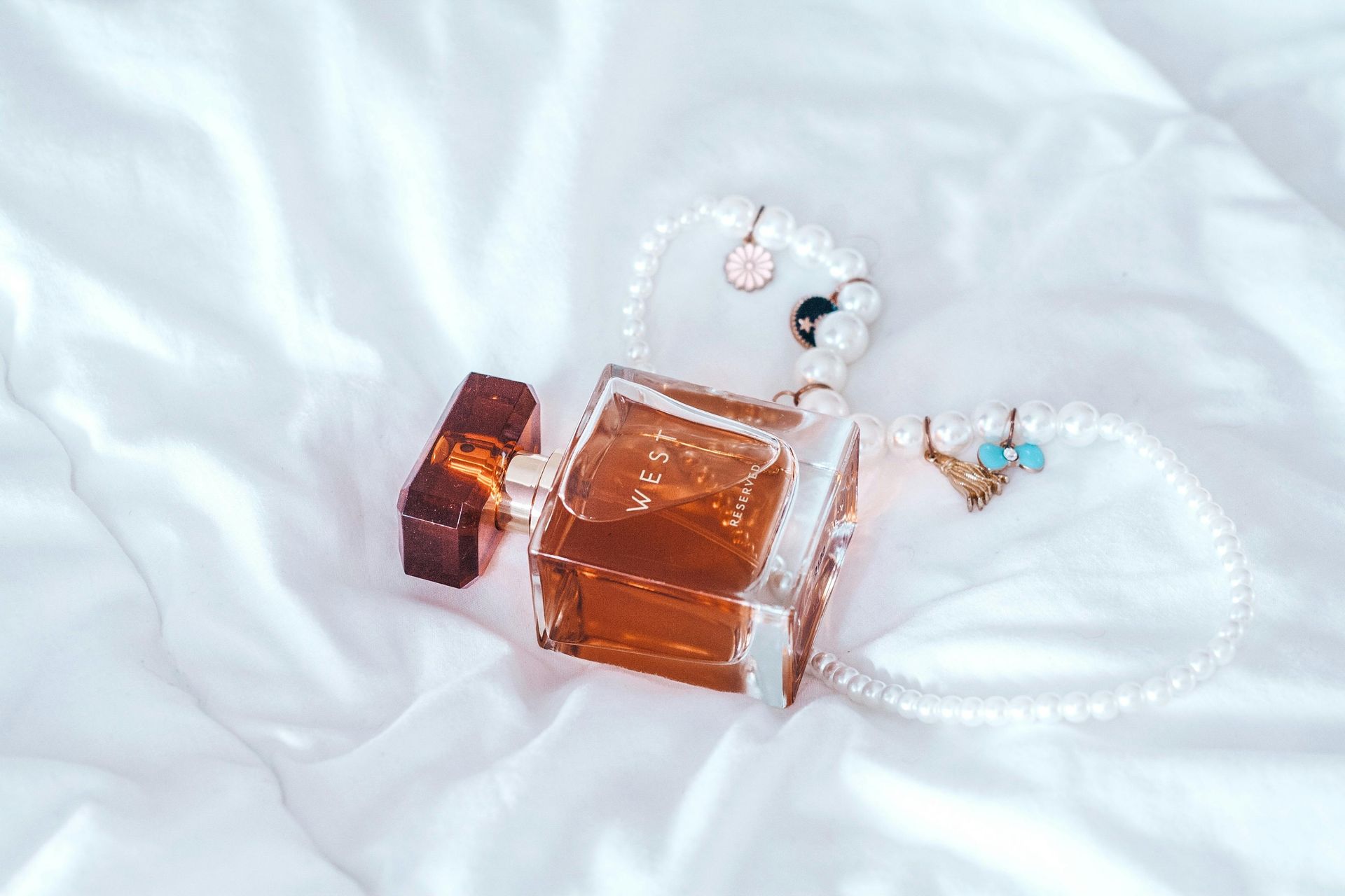 Perfume bottle and pearl jewelry on white satin cloth. The amber-colored perfume bottle is rectangular with a brown cap.