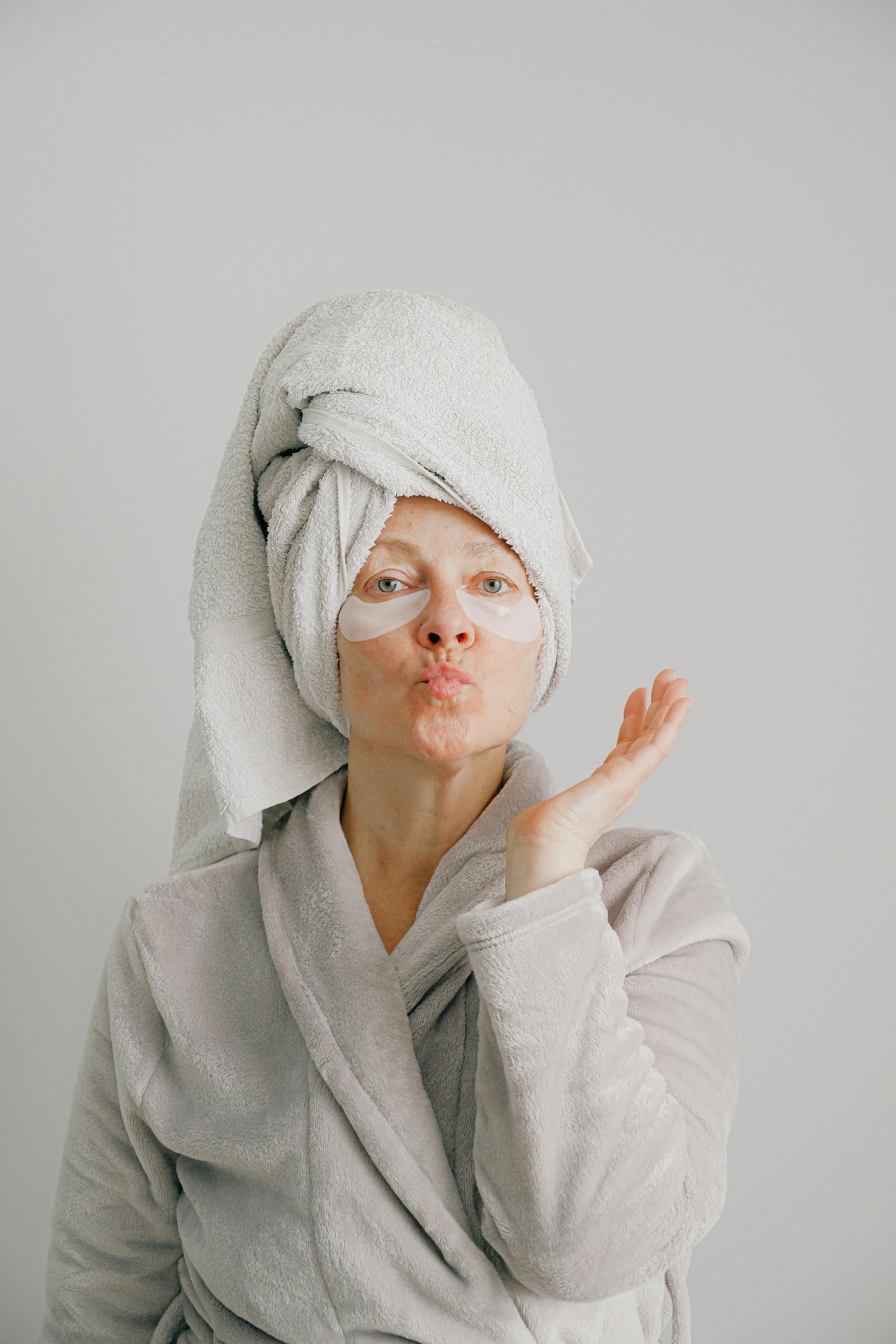 Woman in bathrobe with towel on head and eye patches, blowing a kiss.