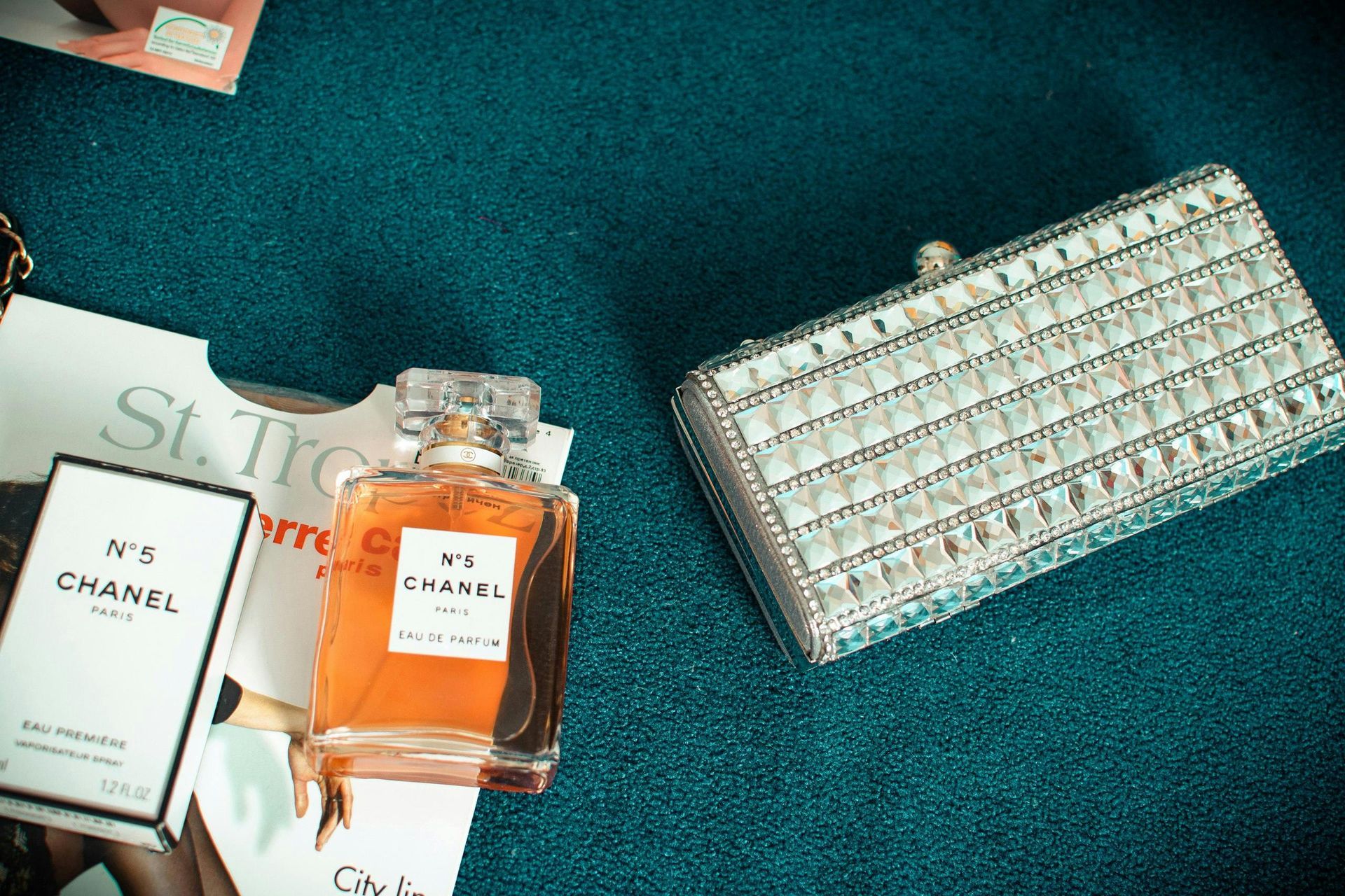 Chanel No. 5 perfume bottle and box beside a jeweled clutch on a teal surface, with parts of a magazine visible.