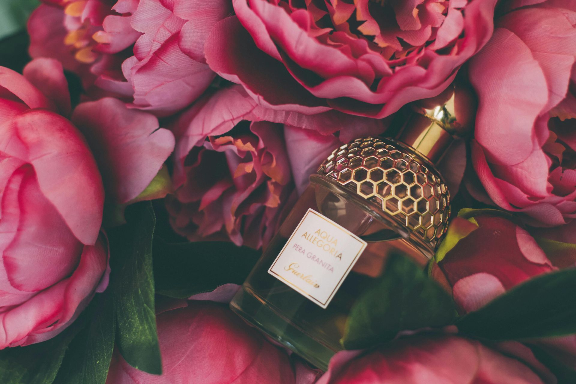A perfume bottle sits among lush pink peonies. The bottle has a honeycomb design and a gold cap.