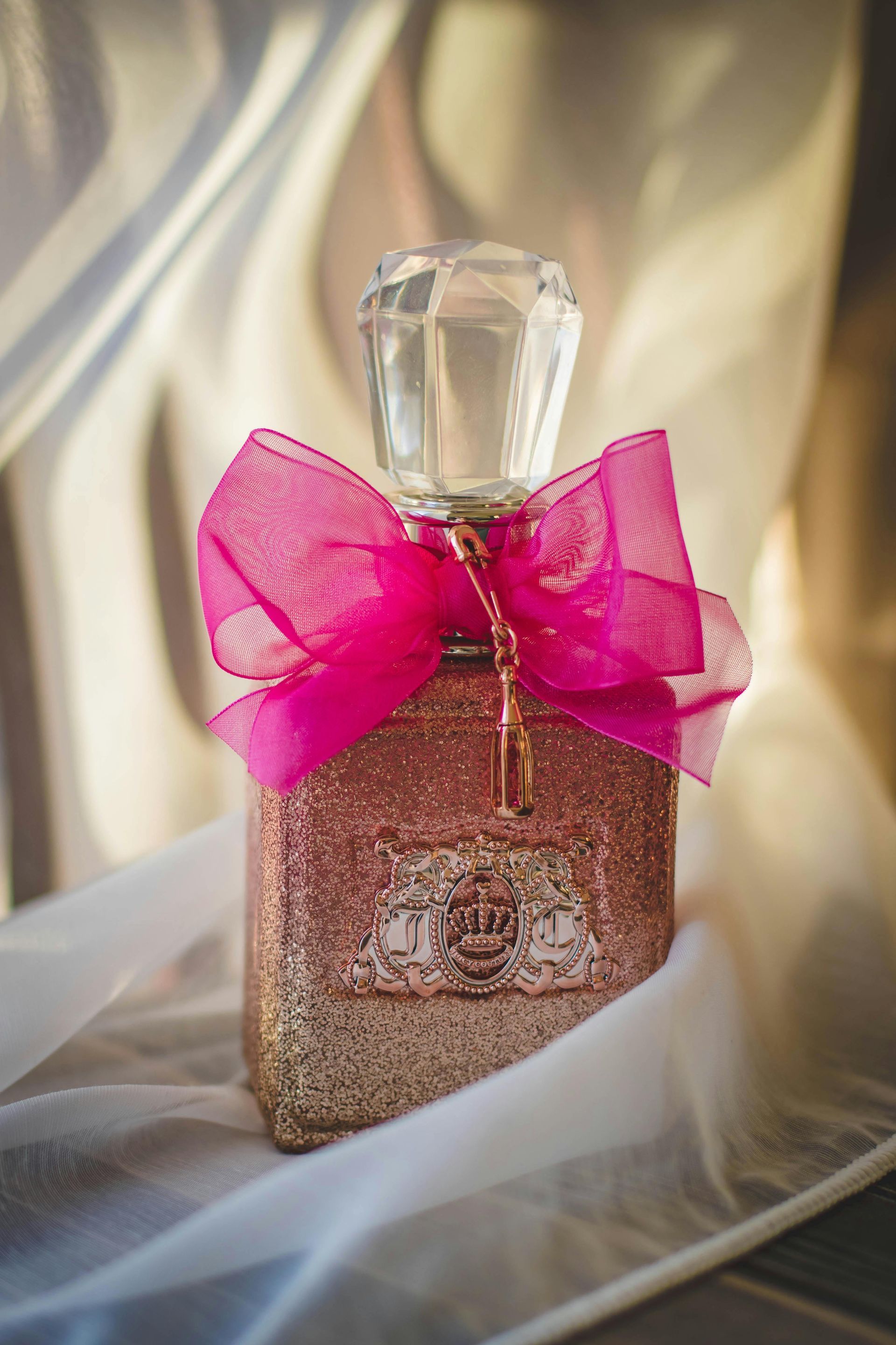 Gold glitter-covered perfume bottle with a faceted crystal top and pink ribbon, set against a white fabric backdrop.