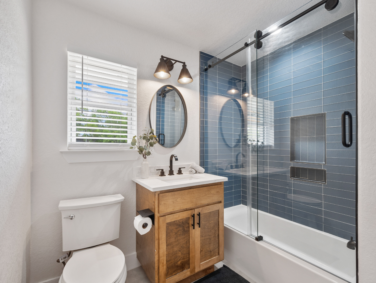 Bathroom with blue tiled shower, wooden vanity, toilet, and window with blinds.