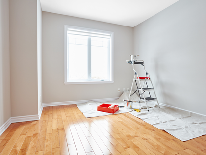 Empty room with hardwood floors being painted; paint supplies and ladder by window.
