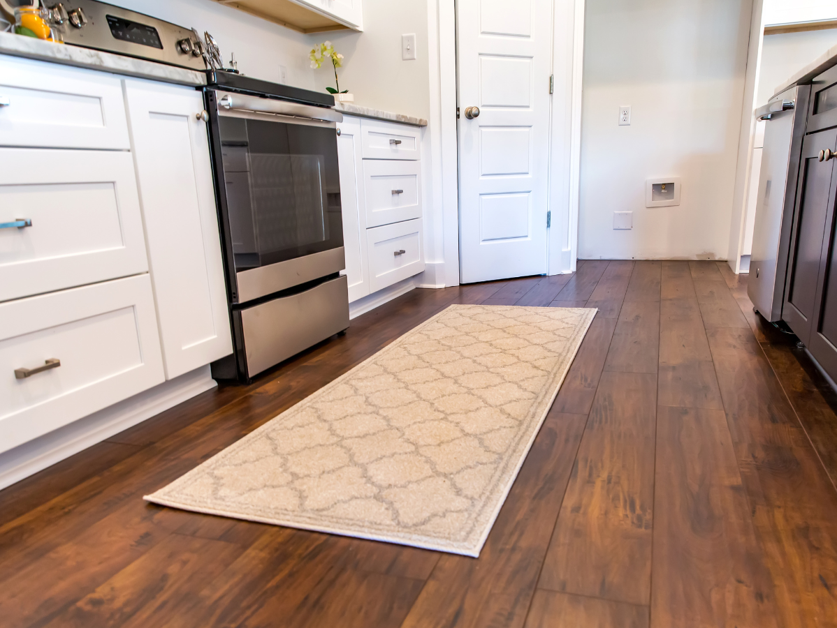 Kitchen with wooden floors, a cream rug, white cabinets, stainless steel appliances, and a closed white door.