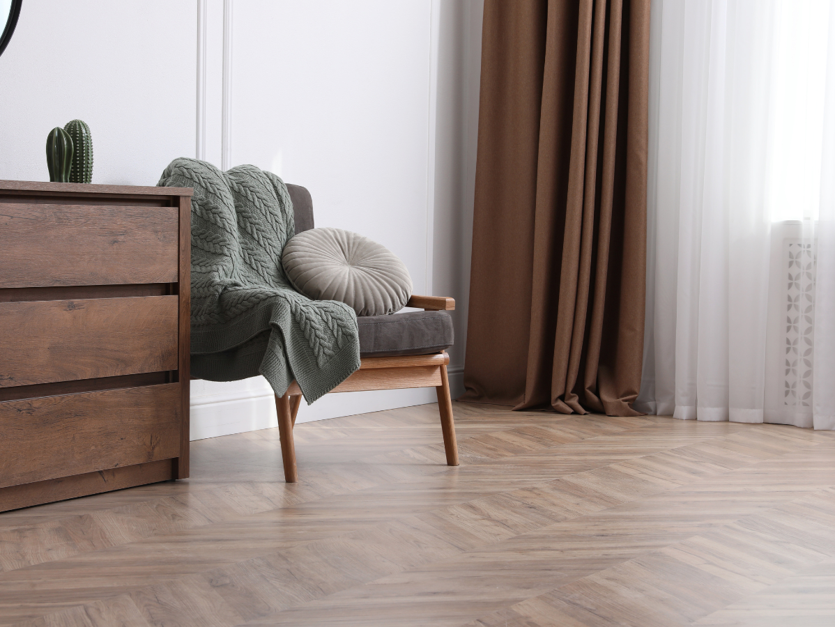 Wooden dresser, gray armchair with green throw and round pillow, brown curtains, wooden floors.