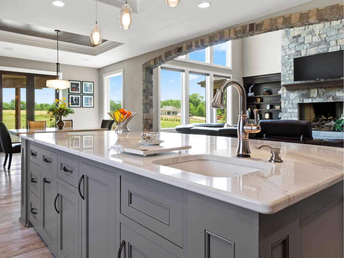 Gray kitchen island with white countertop, sink, and faucet. Dining area and living room in background.