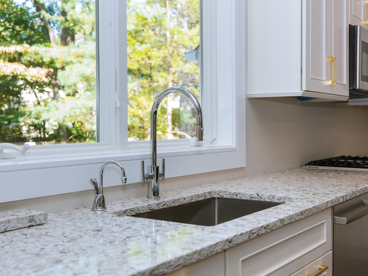 Kitchen sink with stainless steel faucet, white countertops, and a window with a view of trees.