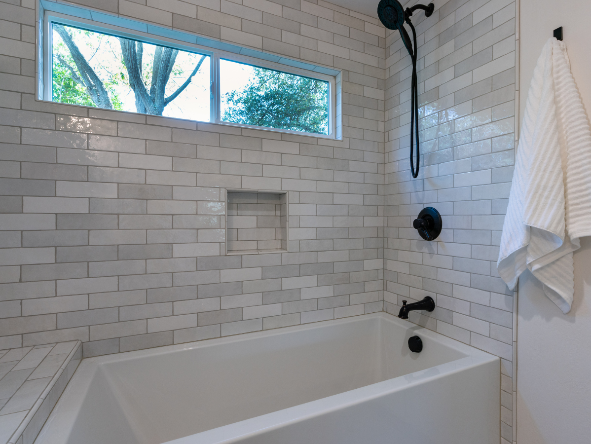 A white tiled bathroom with a bathtub, black fixtures, and a window overlooking trees.