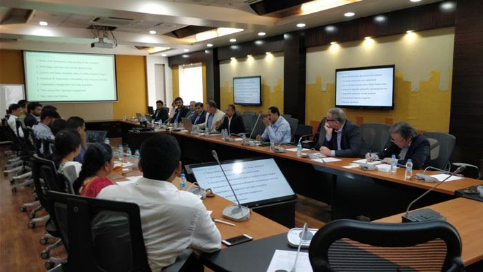 A group of people are sitting at long tables in a conference room.