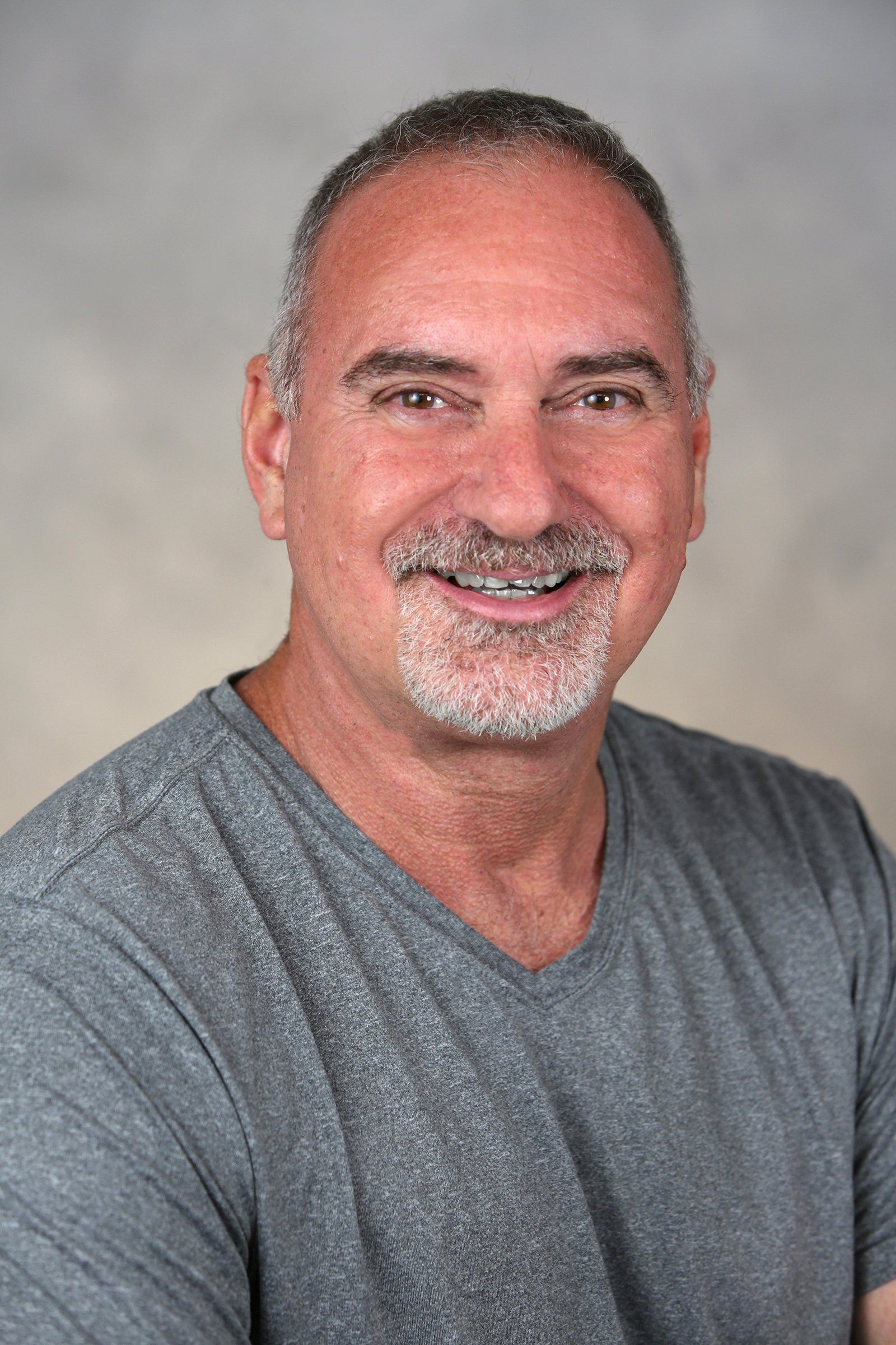 A man with a beard is wearing a grey shirt and smiling for the camera.