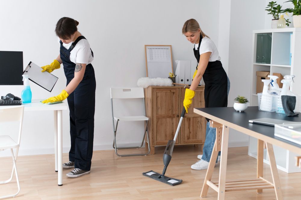 Zwei Reinigungskräfte bei der Arbeit – eine wischt den Boden, die andere reinigt den Tisch im Büro