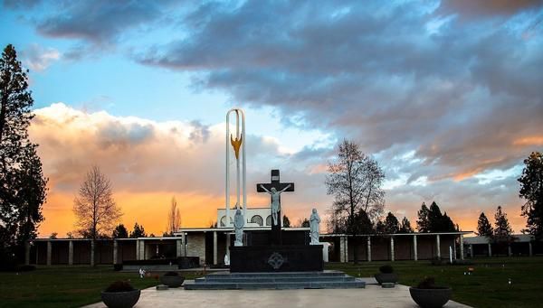 A black crucifix stands in front of a white memorial building under a sunset sky with orange, pink, and blue clouds.