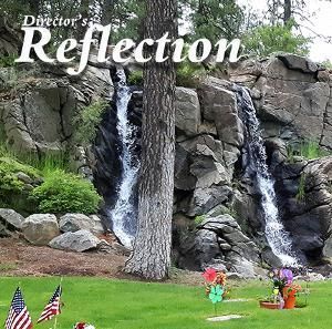 Two waterfalls cascading down a rocky cliff behind a green lawn with American flags and floral tributes.