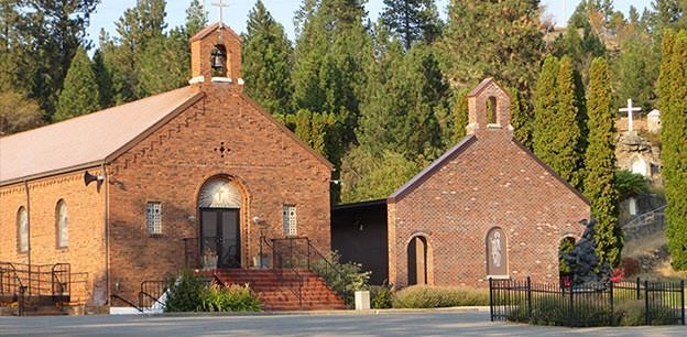 Two brick buildings, a larger chapel with a bell tower and a smaller matching structure, set against a forest backdrop.