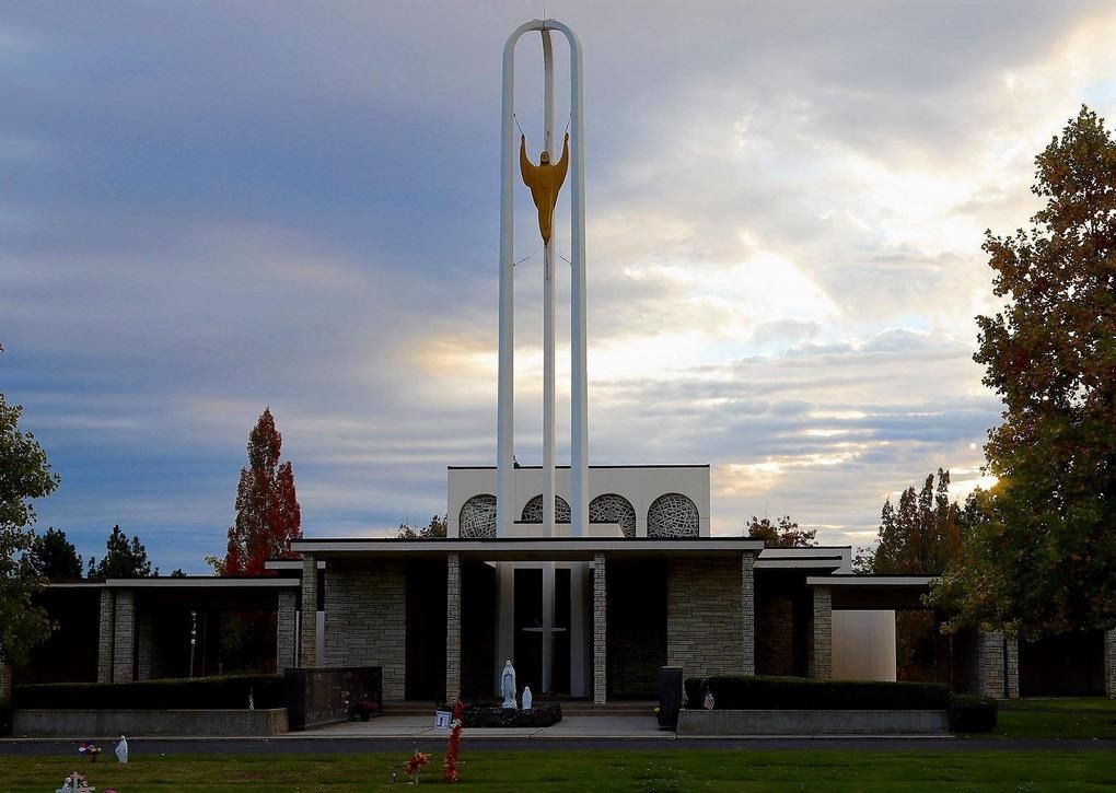Modern cemetery building with stone walls, a tall white spire, and a golden angel sculpture against a twilight sky.