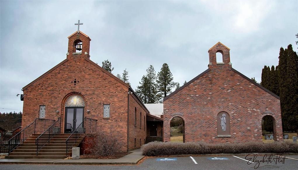 Red brick church building with dual bell towers, a central entrance with steps, and a parking lot in the foreground.