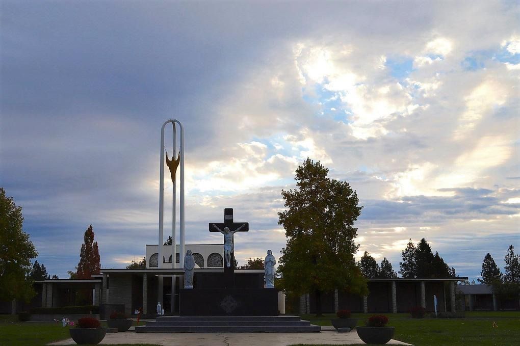 A crucifix and statues stand before a modern church structure with a tall, open-frame tower under a cloudy sky.