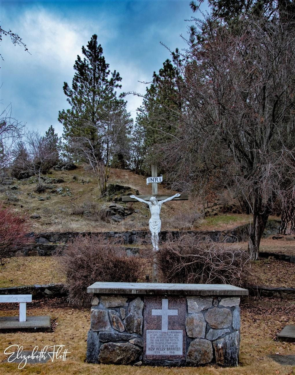 A white crucifix stands on a stone altar base in a peaceful, wooded outdoor setting under a cloudy sky.