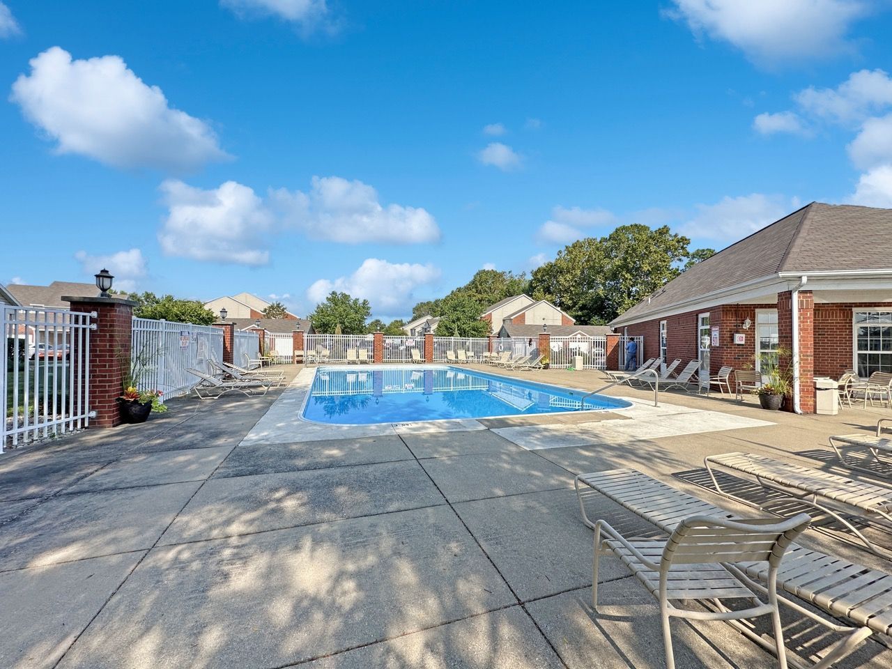 Swimming pool surrounded by lounge chairs and brick buildings under a blue sky with clouds.