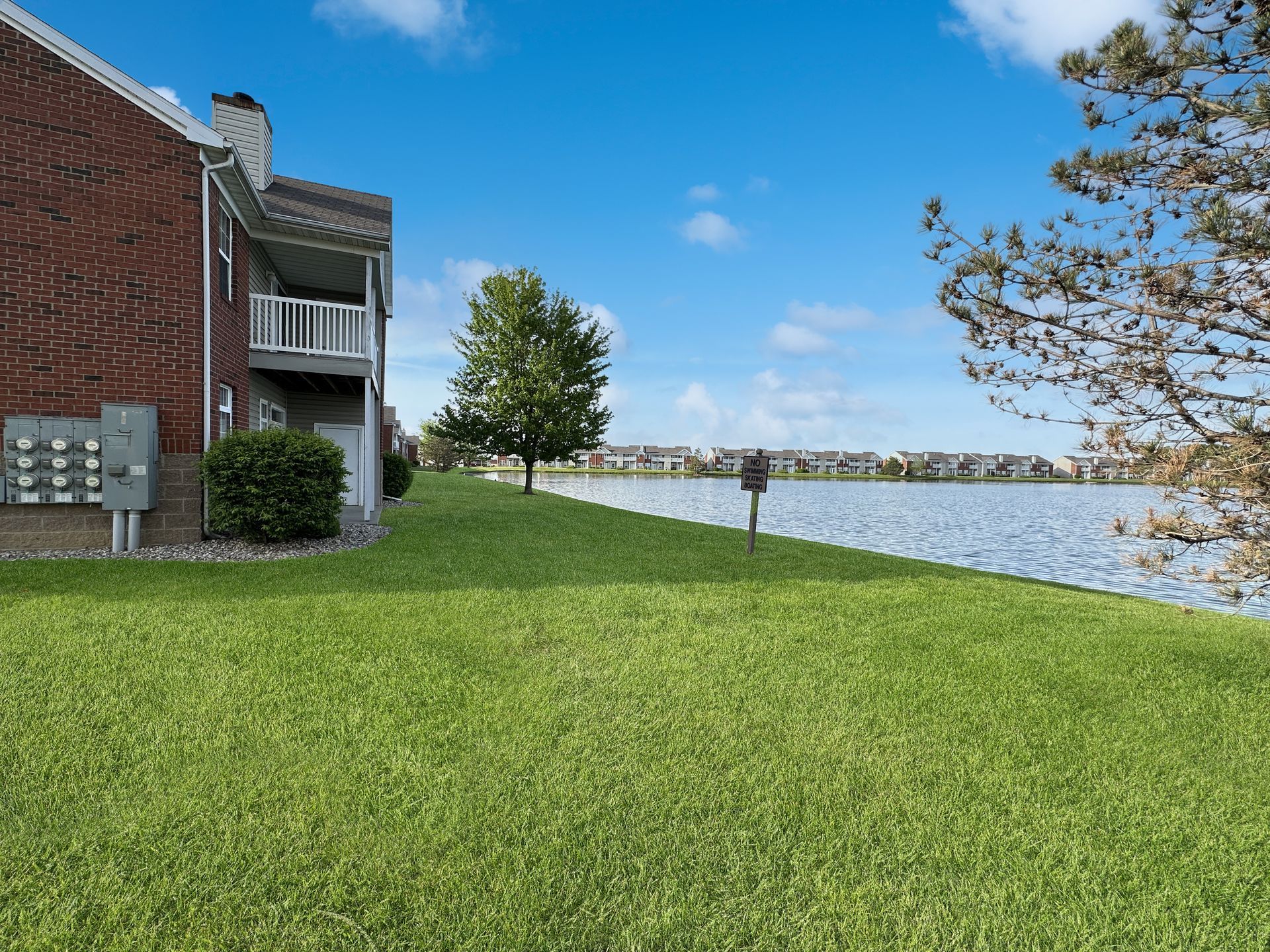 Lakeside view of brick building, green grass, tree, and rippling water under a blue sky.