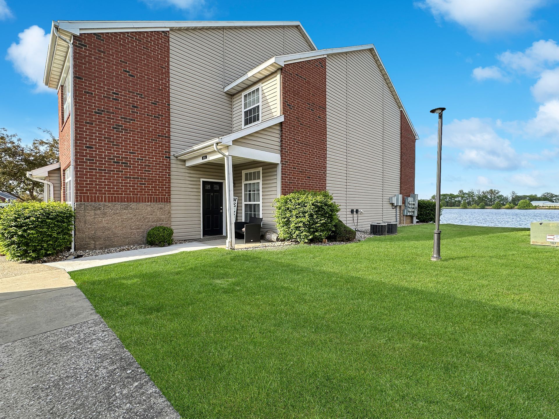 Two-story townhouse with brick accents, green lawn, and a lake in the background under a blue sky.